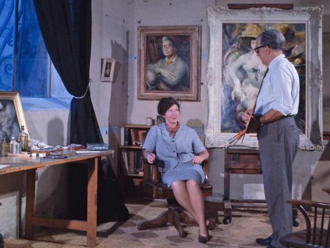 Artists Margaret Olley and William Dobell in Dobell's light filled studio. Olley is seated and looking at Dobell who wears a shirt and tie. They are surrounded by Dobell's paintings.
