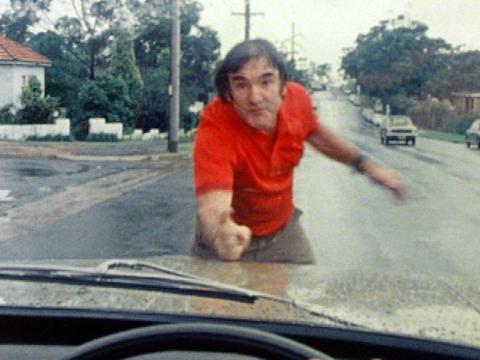 We see a man in a red shirt from the point of view of a car driver who has almost run into him. He is scolding the driver.
