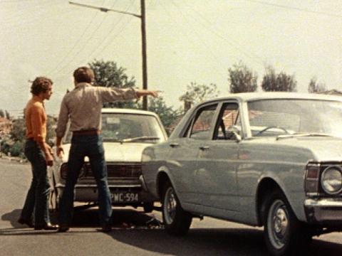 A vintage Falcon and a vintage Cortina car have been involved in a rear-end accident. The drivers stand on the road arguing next to their cars. One of them is pointing his hand.