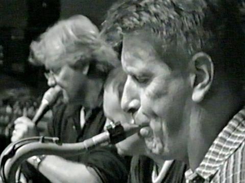 Close up of John Farnham and two other musicians in profile. Farnham is singing into a microphone and the man in the foreground is blowing into a saxophone.