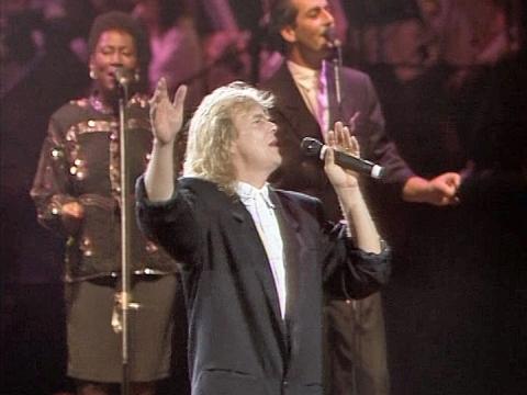 John Farnham singing on stage during a live performance. His backup singers Venetta Fields and Lindsay Field are standing in the background singing.