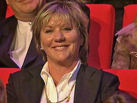 Head and shoulders shot of Jill Farnham, wife of singer John Farnham, smiling, sitting in the audience of a performance by her husband. 