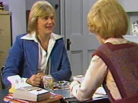 John Farnham in a scene from the TV sitcom Bobby Dazzler (1977), sitting at a desk across from co-star Olivia Hamnett who has her back to the camera.