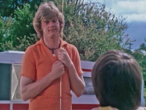 John Farnham, pictured from the waist up, wearing an orange t-shirt and holding a fishing rod is talking to a young boy who has his back to the camera. A red and white caravan can be seen in the background.