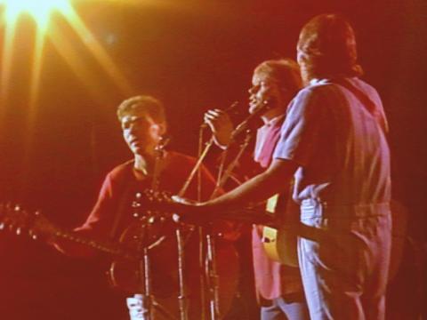 Three members of Little River Band, including John Farnham in the centre, standing on stage behind a microphone. The image has a reddish, yellow glow from the concert lighting.
