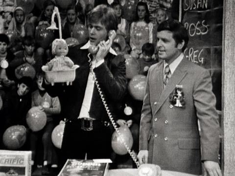 John Farnham and Tony Barber on the set of a TV special for a toy company in 1971. John is holding a toy and speaking into a phone receiver. A studio audience comprised of children, some holding balloons are sitting in the background.
