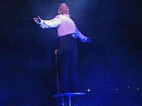 John Farnham on stage during a live production of Jesus Christ Superstar the musical. He is standing on a podium with his arms outstretched and looking upwards.