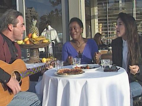 In a casual lunch setting, John Farnham's  longtime backup singers Lisa Edwards, Lindsay Field and Venetta Fields sit around a table. Lindsay is holding an acoustic guitar.