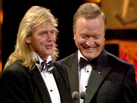 Head and shoulders shot of John Farnham and Bert Newton, both wearing tuxedos and standing in front of a microphone on a stage.