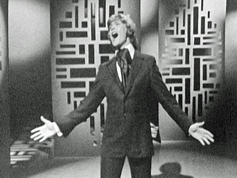 John Farnham, circa early 1970s on the set of a TV show singing with his arms out stretched.