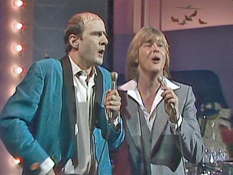Norman Gunston (Garry MacDonald) and John Farnham in a studio setting, both singing into a microphone, circa late 1970s.
