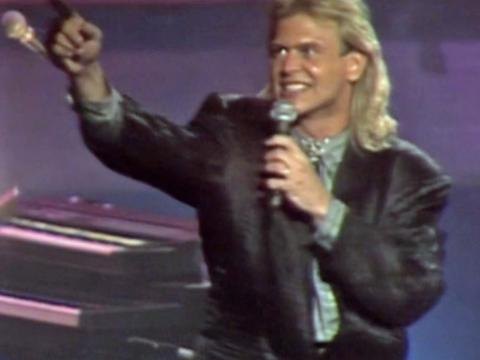 John Farnham on stage singing into a microphone and pointing up towards the audience, circa 1987
