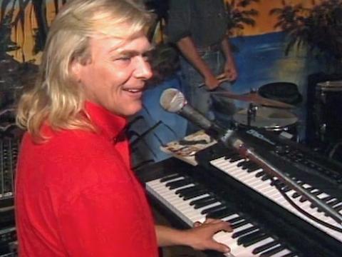 John Farnham sitting in front of a microphone and playing and electric keyboard and looking sideways at his audience