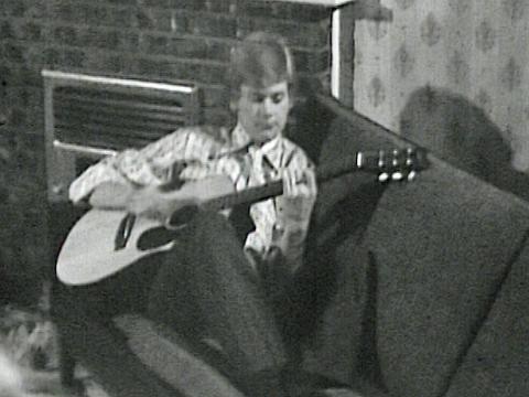 A young John Farnham at home sitting on a couch and strumming a guitar, circa 1970