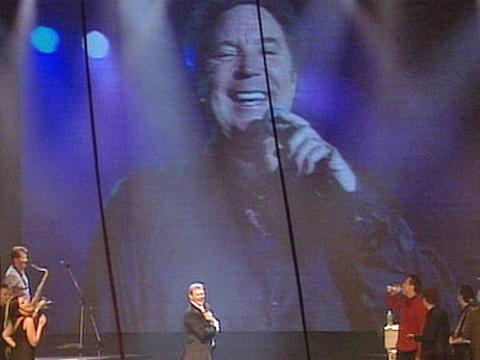 John Farnham on stage during a concert with a large image of Tom Jones behind him on a big screen being broadcast live via satellite.