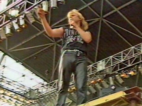 John Farnham on stage, pictured full length from below, dressed in black leather pants and sleeveless shirt and holding a mic in one hand and stretching his other arm out towards the crowd. The stage and lighting rigs can be seen in the background.