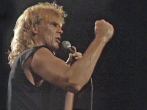 John Farnham in profile from the waist up singing into a mic with one arm raised and hand clenched in a fist.