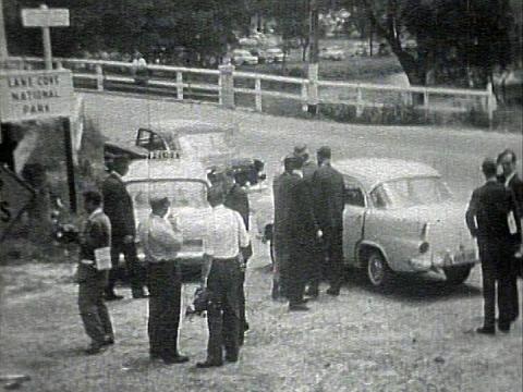 A group of men, likely Police officers and media, standing around cars at one end of Fuller's Bridge in Chatswood, NSW. There is a sign at the end of the bridge that says Lane Cove National Park.