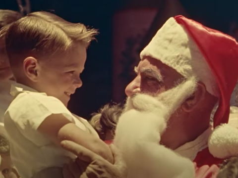 A little boy is being held by a man dressed as Santa. Other smiling children surround them.