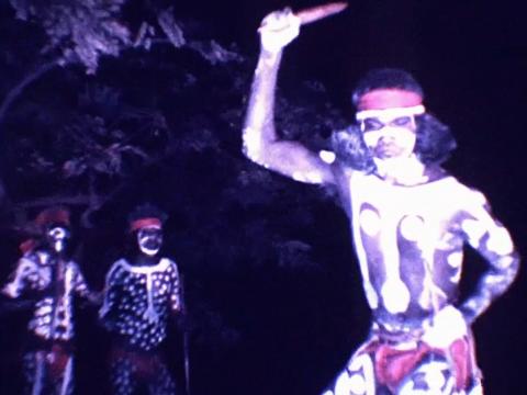  It is night time. A young David Gulpilil wears traditional bodypaint and holds a clap stick above his head as he dances. Two men are in the background, also wearing bodypaint.