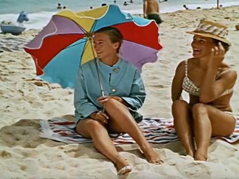 Two young women seated on a striped towel on a Perth Beach, circa 1960. One of the women is wearing a long sleeved top and holding a colourful umbrella, the other woman is wearing a bikini and a hat and both smiling at something off camera to their right.