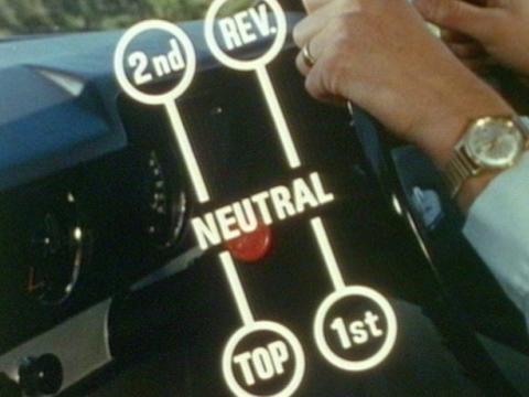 A still of a man with his hands on the steering wheel of a car with the manual gear stick mounted on the steering wheel column. Super imposed over the image is a diagram of the different positions of the stick shift for 2nd, reverse, neutral, top and 1st.