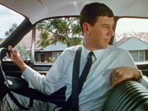 A man in a shirt and tie is reverse parking a vintage car. He is looking behind him to check what's there.