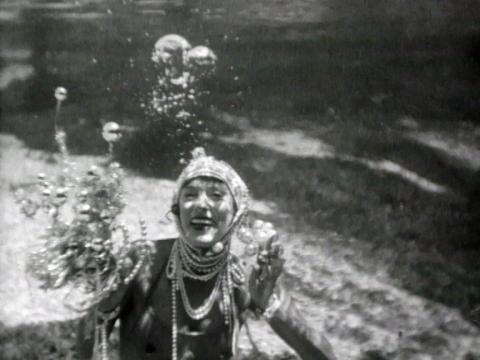 A woman wearing a bathing cap and wearing strings of pearls is underwater with no breathing apparatus. She is looking at the camera.
