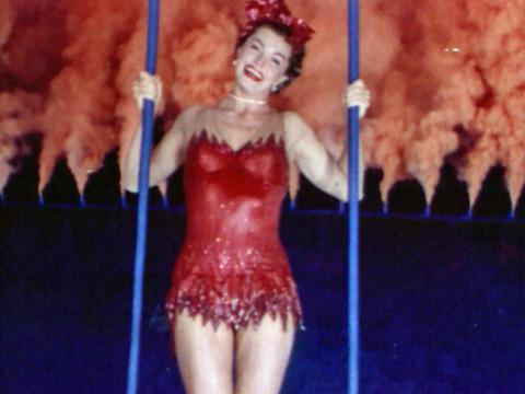 A woman in a red sparkling swimsuit and headwear holds a ring as she is lifted out from a swimming pool. She is surrounded by a circle of men and women in yellow costumes.