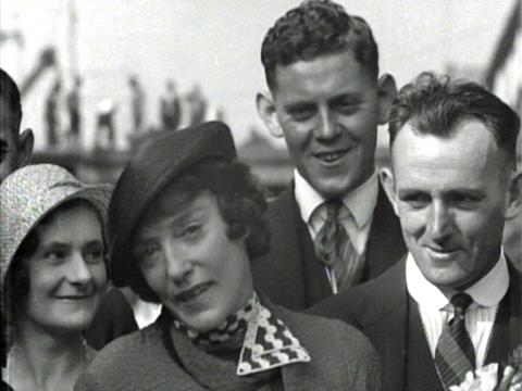 Black and white image of a woman speaking. Three people behind her are listening and reacting to what she says with smiles on their faces.