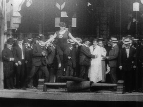 Black and white still of a woman standing on a diving board with her arms outstretched. There is a crowd of men behind her watching.