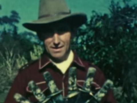 Smoky Dawson wearing hat and holding a fan of throwing knives.