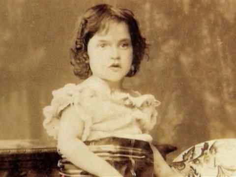 A sepia tinted photograph of a young girl wearing her good outfit for a studio photograph in the late 1800s.