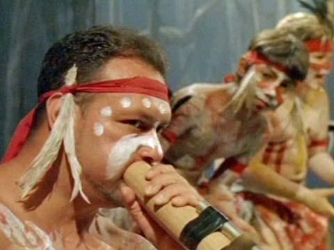 An Aboriginal man in ceremonial dress plays a didjeridu while two boys in ceremonial dress look on