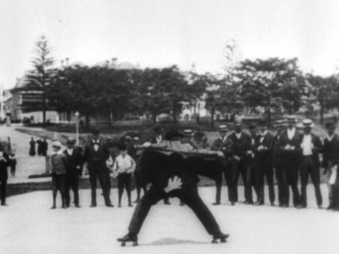 Film still of a man on rollerskates bending over to reveal a white handprint on his backside.