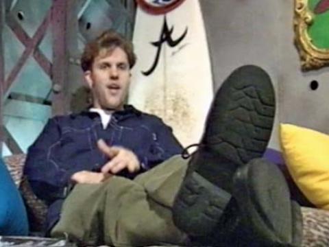 Young man reclining on a bed with a surfboard behind him.