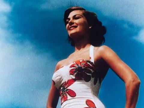 A woman in a bathing suit, pictured from the waist up, the camera angle is looking up towards the sky. She is in a strong pose with her hands on her hips and smiling.