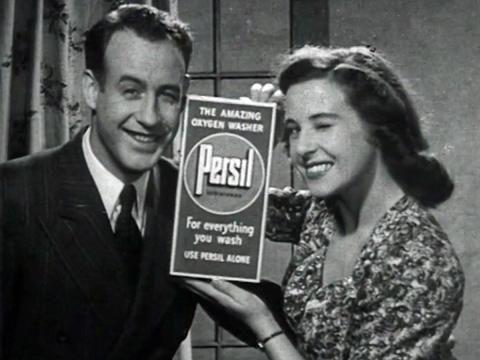 A young man and woman (newlywedded couple) pictured from the waist up smile and wink at the camera. The woman is holding a box of washing powder.