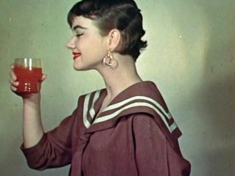 Young woman, standing side-on, holding a glass of orange soft drink and smiling.