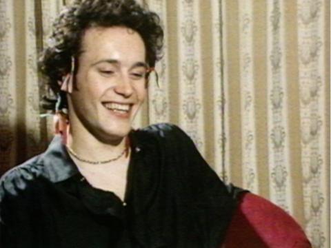Adam Ant, sitting in a hotel suite, laughing as he is interviewed.