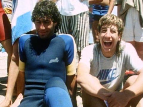 Two men sit in front of the camera with a crowd of people behind them. One is wearing a wet suit the other a Nike t-shirt.