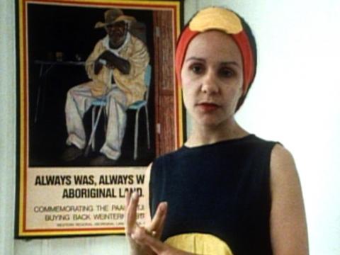 Linda Burney wearing a headband and dress with motifs of the Aboriginal flag in black, red and yellow. In the background there's a poster that says 'Always was, always will be, Aboriginal Land'.