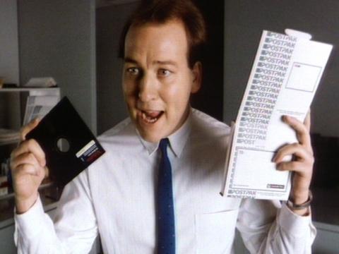 Matt Quartermaine as an office worker wearing a shirt and tie and rapping while he holds a floppy disc and a postpak. 