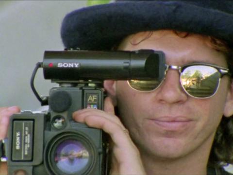 INXS singer Michael Hutchence, wearing reflective sunglasses and a pork pie hat. He is filming with a handicam camera pressed up again his eye.
