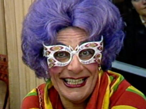 Dame Edna Everage in a purple wig, thoroughly bedazzled winged glasses and a colourful knit.