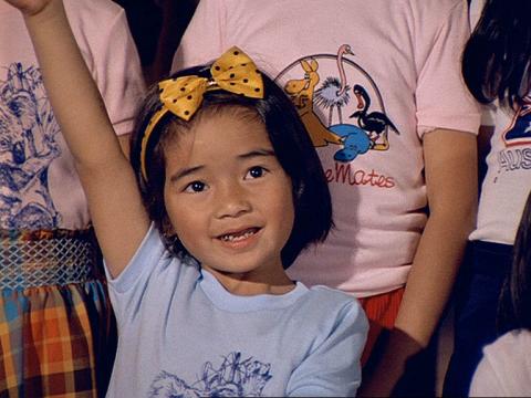 A young girl with a polka dotted yellow bow in her hair has he hand up in the air ready to ask a question.