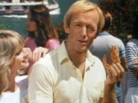 Paul Hogan holds up a prawn to camera while talking. Delvene Delaney looks on lovingly with her hand on his shoulder.