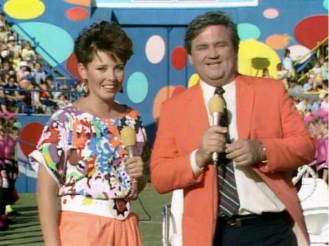Fiona MacDonald and Billy T James in front of a colourful backdrop and crowd. They are both wearing a lot of orange and hold orange microphones.