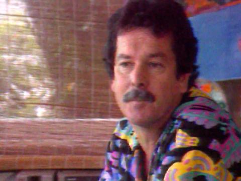 Artist Ken Done sitting in his studio. He's wearing a shirt with one of his colourful designs on it.