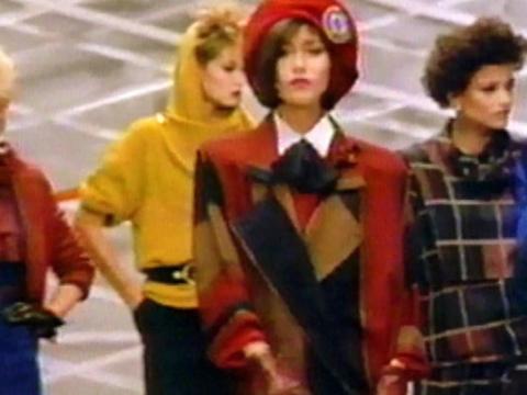 Four models on a catwalk. They are dressed in fashion from the 1980s including spiked hair, hats, shoulder pads and chunky belts.